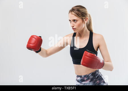 Droit de slim blonde woman 20s'habiller dans les vêtements de sport et des gants de boxe au cours de l'exercice et la forme physique dans le gymnase isolated over white wall Banque D'Images