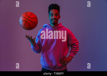 Portrait of african american man masculin à capuche colorés poising à huis clos, avec le basket-ball sur fond violet isolés Banque D'Images
