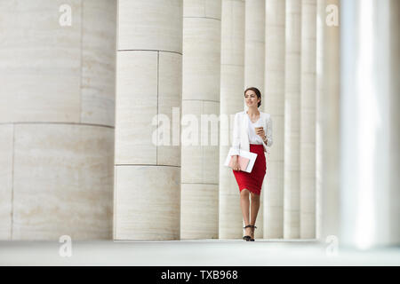 Full Length portrait of beautiful business woman jupe rouge la marche vers la caméra le long de la rangée de piliers, copy space Banque D'Images