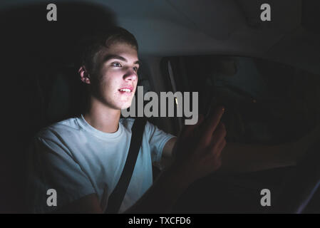 Adolescent distrait au volant d'une voiture avec son téléphone cellulaire dans sa main. La lumière de l'écran du téléphone est éclairant son visage. Banque D'Images