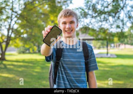 Happy teenager holding son portable tout droit pour afficher un écran vide. Il est debout dans un parc de la ville sur une journée ensoleillée. Banque D'Images