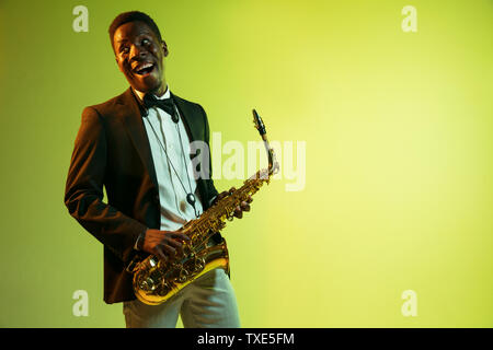 Les jeunes africains-américains musicien de jazz jouant du saxophone sur jaune-vert dégradé fond studio. Concept de la musique, passe-temps, festival. Guy attrayant. improvisation joyeuse Portrait de l'artiste en couleurs. Banque D'Images