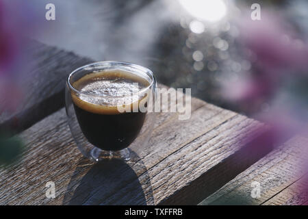 Petit verre double paroi transparente tasse de café frais placé sur une surface en bois naturel, en face d'un étang entouré de fleurs rose floue Banque D'Images