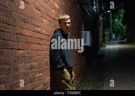 Adolescent à la grave appuyé contre un mur dans une ruelle la nuit. Banque D'Images