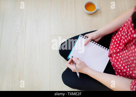 Vue de dessus d'une femme assise sur le sol, qui s'apprête à écrire dans un bloc-notes. Les mains tenant un stylo et un carnet, une tasse de thé à côté. Banque D'Images