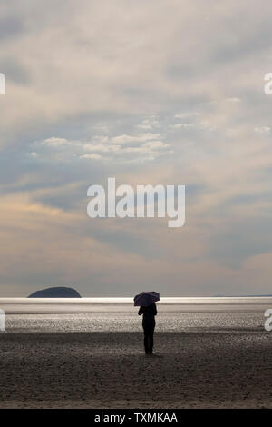 Personnage solitaire distant silhoueté avec parapluie debout isolé sur des sables de plage vides au coucher du soleil, Weston-Super-Mare, Royaume-Uni. Banque D'Images