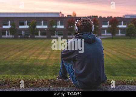 Adolescent assis tout seul en haut d'une colline au coucher du soleil. Il est à l'écoute de la musique à travers son casque. Banque D'Images