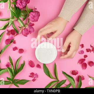 Deux mains d'une jeune fille avec une peau lisse et un pot avec de la crème épaisse et la floraison des pivoines bourgogne avec des feuilles vertes, vue du dessus, concept d'anti-agin Banque D'Images