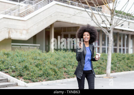 Jeune femme avec des cheveux afro en ville, marchant et parlant au smartphone Banque D'Images
