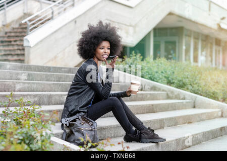 Jeune femme avec des cheveux afro assis sur l'escalier de la ville, parlant à smartphone Blackberry Banque D'Images