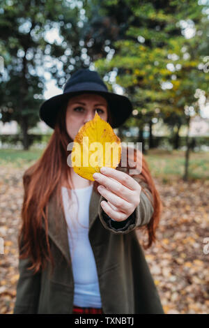 Jeune femme aux longs cheveux rouges holding up autumn leaf in park, portrait Banque D'Images