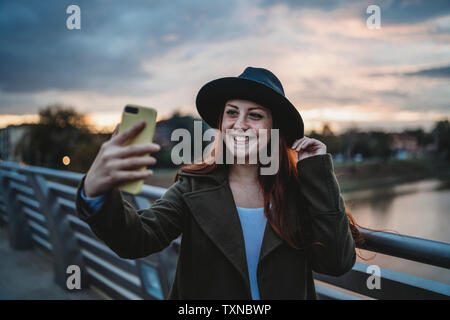 Jeune femme aux longs cheveux rouges sur la passerelle qui selfies smartphone au crépuscule, Florence, Toscane, Italie Banque D'Images