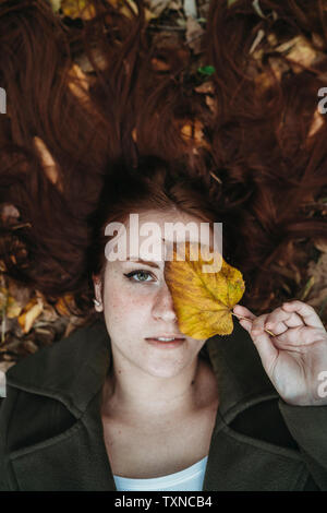 Jeune femme aux longs cheveux rouges se trouvant entre les feuilles d'automne et des feuilles d'automne avec des yeux couvrant les frais généraux, portrait Banque D'Images