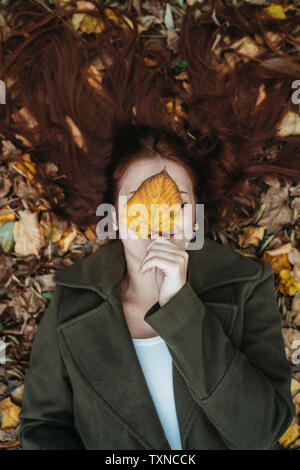 Jeune femme aux longs cheveux rouges se trouvant entre les feuilles d'automne et couvrant le visage avec des feuilles d'automne, d'un portrait Banque D'Images