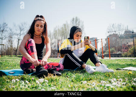 La classe de gymnastique suédoise au sport en plein air, les jeunes femmes sitting on grass lacets de liage et looking at smartphone