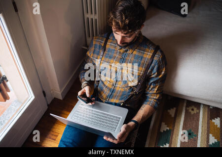 Mid adult man sitting on floor with laptop salon using smartphone Banque D'Images