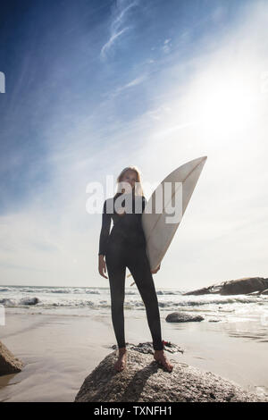 Jeune femme surfer carrying surfboard on beach rock, pleine longueur portrait, Cape Town, Western Cape, Afrique du Sud Banque D'Images