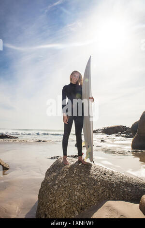 Jeune femme surfer carrying surfboard on beach rock, pleine longueur portrait, Cape Town, Western Cape, Afrique du Sud Banque D'Images