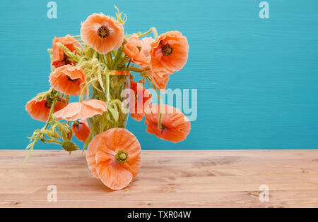 De nombreuses et belles roses coupées avec de longues tiges sinueuses coquelicots, débordement d'un vase en verre sur une table en bois, contre un fond bleu turquoise peint Banque D'Images