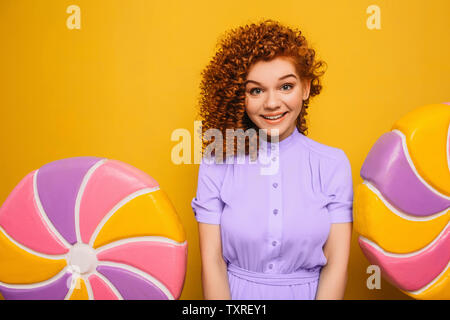 Rousse bouclée femme positive portant robe bleue avec sweet big lollipop sur fond jaune Banque D'Images