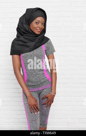 Jeune femme musulmane élégante en noir portant le hijab et gris sportswear posant contre un mur de briques blanches. Beau, positif, slim modèle africain debout, looking at camera, souriant. Banque D'Images