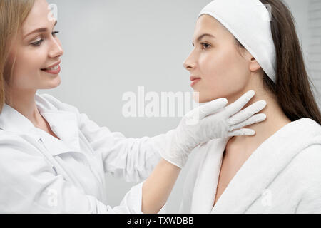 Jeune, jolie femme sur les conseils du cabinet en cosmétologie. Médecin positive face à l'inspection professionnelle du patient, le toucher avec les mains en blanc gants médicaux. Cosmetologist et client sur fond gris. Banque D'Images