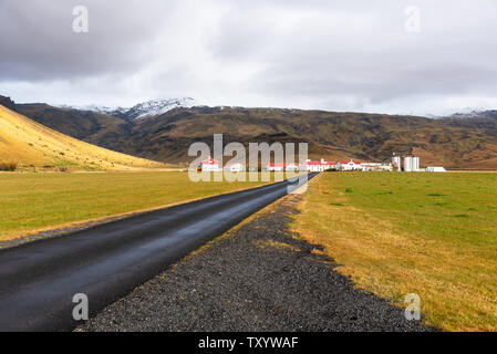 Route pavée vide vers une ferme au pied de la montagne dans l'Islande sur un jour d'automne nuageux Banque D'Images