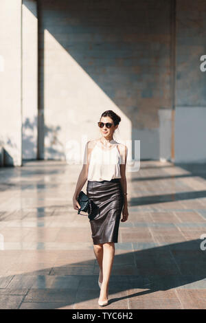 Business Woman vêtu d'un cuir jupe en mousseline de soie, ou chemisier, porter des chaussures à talons et lunettes de marcher près d'un bâtiment. Jeu de lumière Banque D'Images