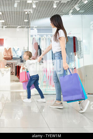 Vue latérale du jeune belle mère shopping avec ma petite fille dans centre commercial. Fille positive de maintien et d'exécution dans les magasins. Famille le choix et l'achat de nouveaux vêtements et des jouets. Banque D'Images