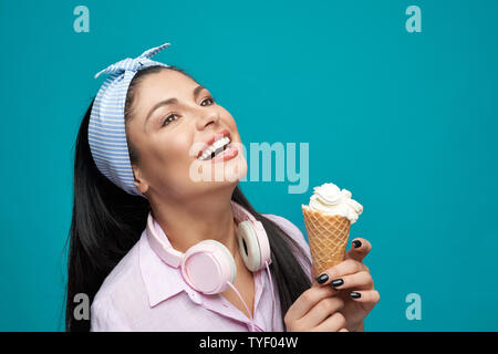 Focus sélectif de jeune brunette élégant gardant la crème glacée et de rire sur fond isolé bleu. Jolie fille à côté, posant et profiter de l'heure d'été. Concept de se détendre et se reposer. Banque D'Images