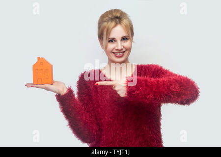 Vous voulez propre maison ? Jeune femme de l'agent immobilier en rouge blouse debout, tenant du papier jaune petite maison et vous proposons de la planification de nouvelles home à acheter. Ind Banque D'Images