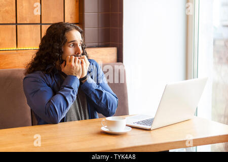 Portrait de peur beau jeune homme adulte freelancer dans style casual assis dans un café et regarder des horreurs film dans coffre, mordent les ongles, bussinessman Banque D'Images
