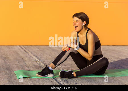 Jeune fille sportive en noir sportwear assis sur mat et ont un fort mal problème avec genou, spasme douloureux. La jambe de préhension avec la main et crier. Out Banque D'Images