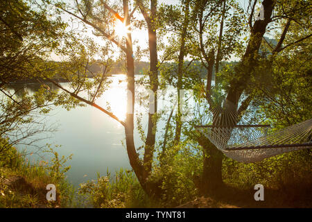 Un coton hamac suspendu entre deux arbres surplombant un lac et le soleil est un sommet des arbres jeter Banque D'Images