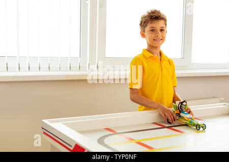 Sourire heureux. Positive joyeux garçon debout à la table tout en tenant sa petite voiture Banque D'Images