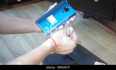 Man holding Huawei Mate 20 X bleu smartphone avec une main et enlever ruban de protection de sa façade arrière au-dessus de sa boîte noire ouverte. Color gradi Banque D'Images