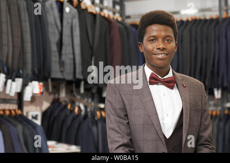 Portrait of smiling handsome african man en chemise blanche, gilet noir, veste gris, noeud papillon rad pose, looking at camera. Grand choix de Costumes et vestes sur des cintres sur arrière-plan. Banque D'Images