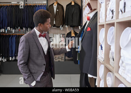 Jeune homme africain choisissant costume élégant à la mode en boutique. Client debout près de mannequin en costume et à la recherche à veste. Grand choix de chemises blanches sur des étagères et des vestes sur des supports en ligne. Banque D'Images