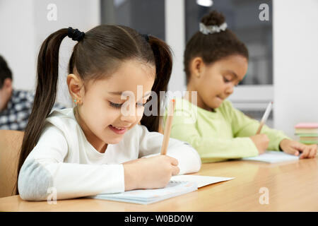 Jolie, heureuse de l'école primaire les écolières sitting at desk in classroom, holding crayons et soigneusement écrit en cahier. Les filles à la recherche dans l'ordinateur portable, ce qui pose, en souriant. Banque D'Images