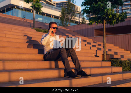 Freelancer moderne talking on smartphone et à l'aide de laptop while sitting on steps au coucher du soleil. Banque D'Images