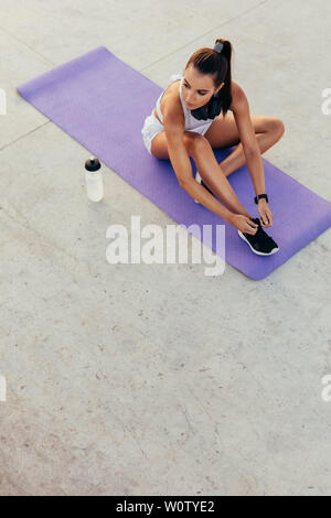 Femme assise sur un tapis d'exercice attachant ses lacets et regardant loin. femme de fitness se préparant pour l'entraînement de yoga. Banque D'Images