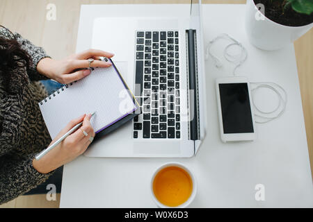 Vue de dessus d'une jeune femme assise à la table à la maison en face de l'ordinateur portable, de prendre des notes dans l'ordinateur portable. Tasse de thé et un smartphone à côté. Banque D'Images