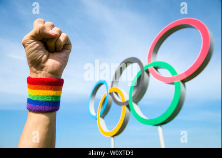 Londres - 4 mai 2019 : une main de couleur arc-en-ciel de la fierté de porter le bracelet de LGBTQI fait un poing de célébration en face d'Anneaux olympiques en bleu ciel. Banque D'Images