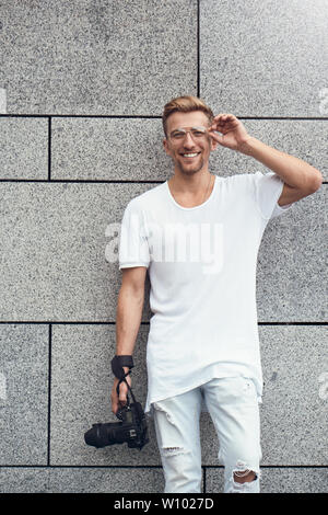 Photo de handsome guy Caucasienne sur mur texturé gris en blanc T-shirt et jeans blanc avec un appareil photo accroché sur la sangle sur son bras. Banque D'Images