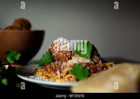 Gros plan d'une assiette de spaghetti et boulettes de viande rustiques avec un bol de boulettes de viande, de fromage parméséen et de persil frais. Banque D'Images