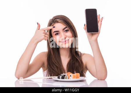 Smiling young asian woman eating sushi à la table isolated over white background, montrant écran blanc téléphone mobile Banque D'Images