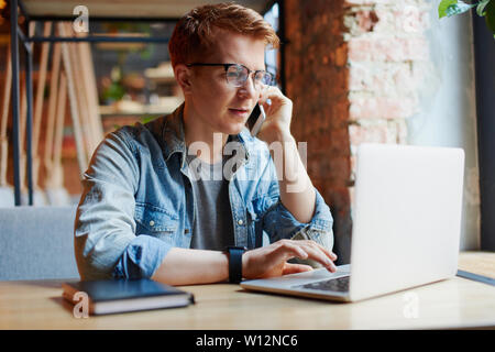 L'homme en chemise en jean types sur l'ordinateur portable et parle au téléphone. Banque D'Images