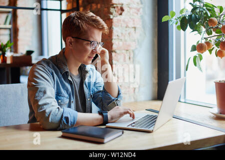 L'homme en chemise en jean types sur l'ordinateur portable et parle au téléphone. Banque D'Images