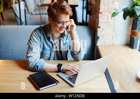 L'homme en chemise en jean types sur l'ordinateur portable et parle au téléphone. Banque D'Images