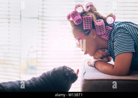 L'amour en moment cocasse avec femme s'étendit sur le canapé avec des bigoudis rose sur des cheveux noir et jolie chien pug l'embrasser ou faire de belle expression ongue - Banque D'Images